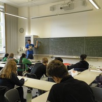 Ein Klassenraum ist zu sehen. Gefüllt ist er mit rund 10 sitzenden Jugendlichen. An der Front des Klassenraums steht Daniel Timms und zeigt auf eine Schultafel. Auf dieser Tafel sind vielstellige Zahlen zu sehen, die miteinander multipliziert werden. 