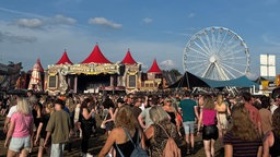 Ein Blick über ein Festivalgelände, mit vielen Menschen, einer Bühne und Riesenrädern