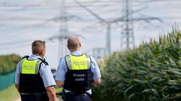 Zwei Polizisten stehen in einem Feld und blicken auf Strommasten im Hintergrund