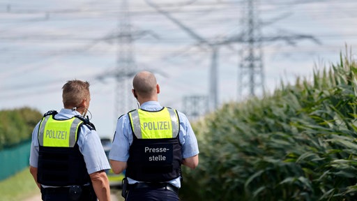 Zwei Polizisten stehen in einem Feld und blicken auf Strommasten im Hintergrund
