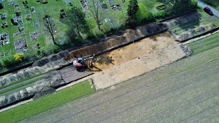 Luftaufnahme einer Ausgrabungsstätte an einem Friedhofsrand in Frotheim. Ein Bagger und mehrere Personen arbeiten in einem freigelegten, rechteckigen Areal mit heller Erde.