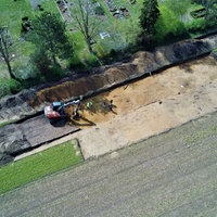 Luftaufnahme einer Ausgrabungsstätte an einem Friedhofsrand in Frotheim. Ein Bagger und mehrere Personen arbeiten in einem freigelegten, rechteckigen Areal mit heller Erde.