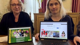 Zwei Frauen sitzen mit zwei Laptops, auf denen sie zwei Internetseiten präsentieren, an einem Tisch