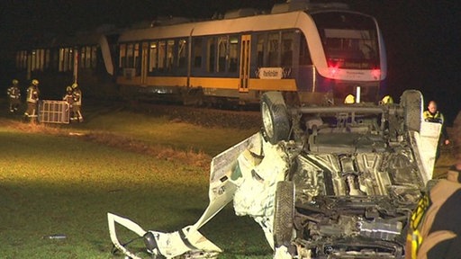 Ein demoliertes Auto liegt auf einer Wiese auf dem Dach. Im Hintergrund sind Rettungskräfte und ein Zug zu sehen. 