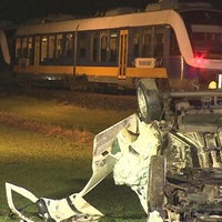 Ein demoliertes Auto liegt auf einer Wiese auf dem Dach. Im Hintergrund sind Rettungskräfte und ein Zug zu sehen. 