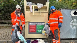 Zu sehen ist ein voller Altkleidercontainer und zwei Mitarbeiter der Stadt Düren in Arbeitskleidung.
