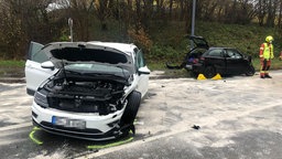 Weißes zerbeultes Auto im Vordergrund. Straße mit weißem Löschschaum bedeckt. Im Hintergrund ein verunfalltes schwarzes Auto und ein Feuerwehrmann.