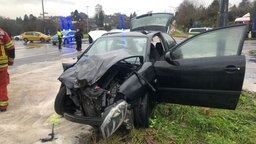 Schwarzes Auto vorne zerbeult. Türen stehen auf. Im Hintergrund zwei Polizeiautos und Polizisten
