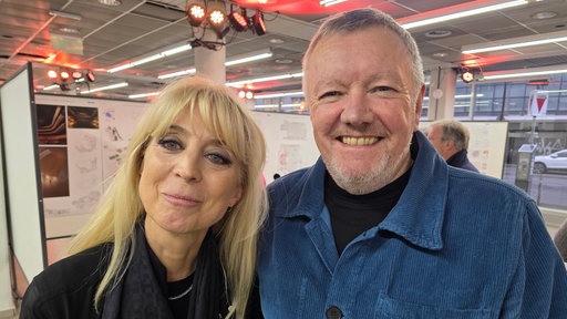 Ein älterer Mann mit Drei-Tage-Bart und eine blonde Frau lächeln vor einer Reihe von Stellwänden