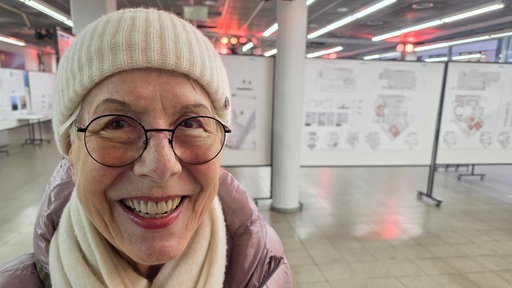 Eine ältere Frau mit runder Brille, weißer Mütze, weißem Schal und dicker Jacke vor einigen Stellwänden