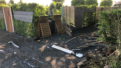 Kaputte Holzverkleidung und verstreute Trümmer auf einem Kiesweg abseits eines Gartnens. 