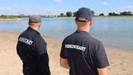 Zwei Männer mit Kappe und Ordnungsamt-Jacken an einem Rheinstrand