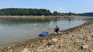Eine Frau sitzt am Ufer der Bevertalsperre. Neben ihr schwimmt eine Luftmatratze. Zwei Kinder schwimmen im Wasser