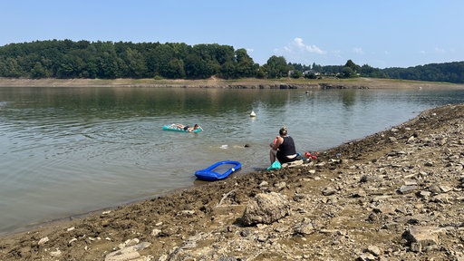 Eine Frau sitzt am Ufer der Bevertalsperre. Neben ihr schwimmt eine Luftmatratze. Zwei Kinder schwimmen im Wasser