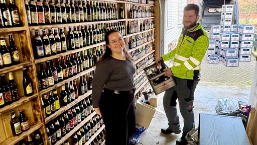 Ein Mann und eine Frau stehen in einer Garage, deren Wände voller Bier sind, im Hintergrund sieht man viele Bierkisten.