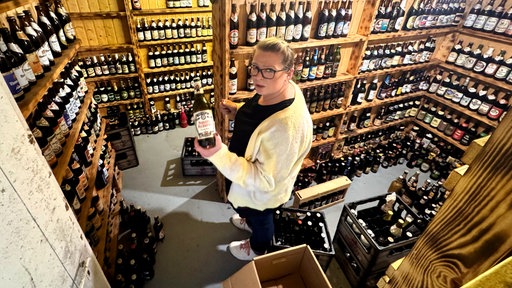 Frau steht im Keller voller Bier und hält eine Flasche in der Hand. 