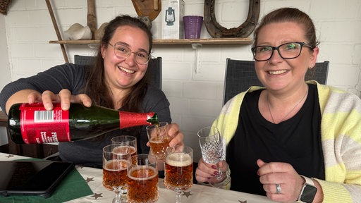 Zwei Frauen sitzen am Tisch und gießen bräunliches Bier in Gläser.