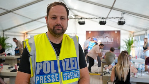 Ein Mann mit dunklen Haaren und Drei-Tage-Bart steht in einem großen Zelt mit vielen Menschen im Hintergrund. Er trägt eine leuchtend gelbe Weste mit der Aufschrift "Polizeipressesprecher"