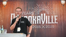 Ein Mann mit Brille, der vor einem Rednerpult steht und ein Mikrofon vor dem Mund hat. Hinter ihm an der Wand steht geschrieben: "10 Years Parookaville 2015 X 2025"