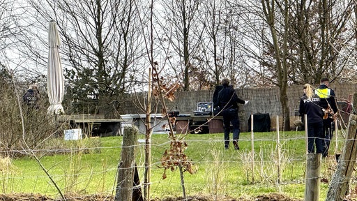Hinter einem Maschendrahtzaun befinden sich drei Personen, davon zwei Polizisten mit Warnweste, auf einem grünen Feld.