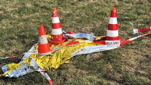 Rot-weiß gestreifte Pylone stehen auf einer grünen Wiese, davor Flatterband mit der Aufschrift "Polizei".