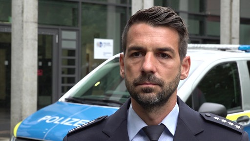 Ein Polizist, Simon Rott, steht vor einem Polizeiauto