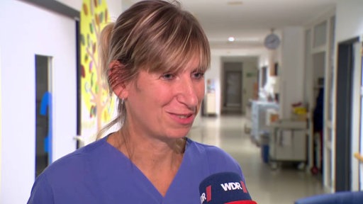 Eine Frau in Arztkleidung steht in einem Flur und gibt ein Interview