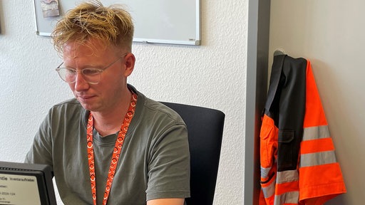 Ein Mann arbeitet am Computer, im Hintergrund eine Jacke in Signalfarben
