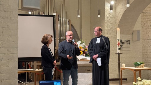 Eine Frau steht links neben zwei Männern in einer Kirche. der Mann in der Mitte hält ein Mikrofon. 