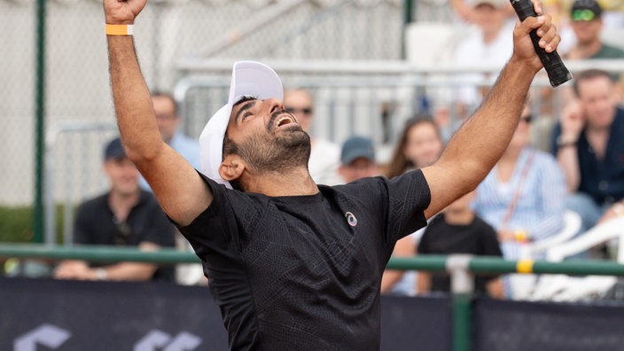 Mika "Tenniszons" steht auf dem Tennisplatz, hebt die Arme und den Kopf nach oben und freut sich