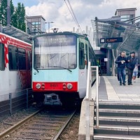 Zwei Stadtbahnen der Stadtwerke Bonn stehen auf den Gleisen der Haltestelle Stadthaus. Auf dem Bahnsteig laufen Menschen.