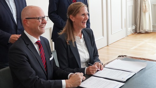 Ein Mann und eine Frau sitzen nebeneinander an einemTisch und lachen. Vor ihnen liegt ein Vertrag und die Frau hält einen Stift in der Hand.