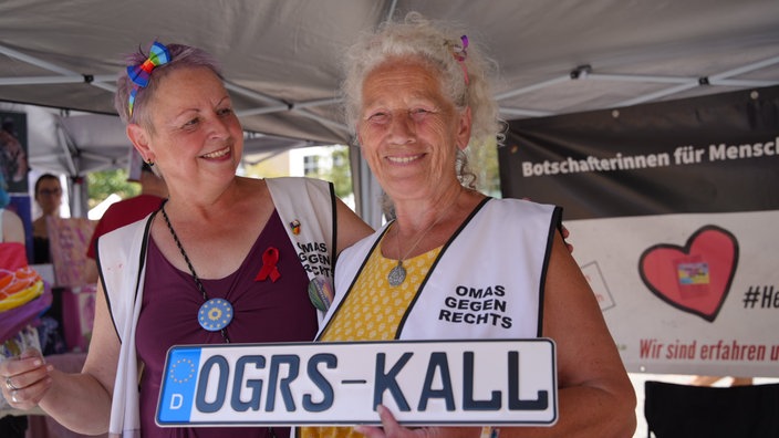 Zwei Frauen von den Omas gegen Rechts tragen weiße Westen und halten ein Nummernschild ain den Händen.