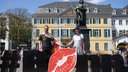 Sebastian hat sich eine Regenbogenfahne umgebunden und hält Sarahs Hand. Vor ihnen der Schriftzug "Bonn". Das o ist ein Kussmund.