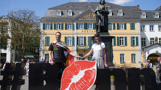 Sebastian hat sich eine Regenbogenfahne umgebunden und hält Sarahs Hand. Vor ihnen der Schriftzug "Bonn". Das o ist ein Kussmund.