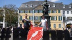 Sebastian hat sich eine Regenbogenfahne umgebunden und hält Sarahs Hand. Vor ihnen der Schriftzug "Bonn". Das o ist ein Kussmund.