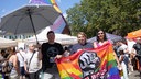 CSD_Teilnehmer Sascha hält einen schwarzen Schirm in der rechten Hand, in der linken Hand hält er mit Dani und einer dritten Person eine Regenbogenfahne.