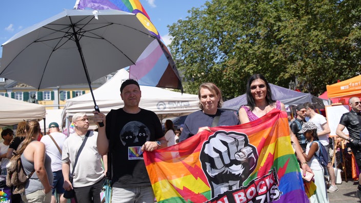CSD_Teilnehmer Sascha hält einen schwarzen Schirm in der rechten Hand, in der linken Hand hält er mit Dani und einer dritten Person eine Regenbogenfahne.