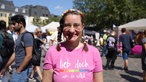 Eine Frau mit einem pinken T-Shirt und Brille steht auf einem Markplatz. Sie schaut lächelnd in die Kamera. Auf dem T-Shirt steht "Lieb doch, wen du willst".