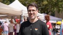 Ein Mann mit einem schwarzen T-Shirt und einem Regenbogenherz darauf schaut lächelnd in die Kamera. Im Hintergrund stehen mehrere Pavillons auf einem Marktplatz.