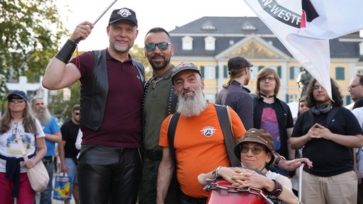 Vier Menschen im Portraitfoto lachen in die Kamera. Einer hält eine Flagge, eine andere Person sitzt in einem Rennauto-ähnlichen Rollstuhl