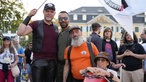 Vier Menschen im Portraitfoto lachen in die Kamera. Einer hält eine Flagge, eine andere Person sitzt in einem Rennauto-ähnlichen Rollstuhl