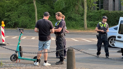 Ein Polizist in Uniform steht neben einem Mann in schwarzem Shirt. Danaben steht ein E-Sooter. Es ist eine Kontrolle.