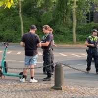 Ein Polizist in Uniform steht neben einem Mann in schwarzem Shirt. Danaben steht ein E-Sooter. Es ist eine Kontrolle.