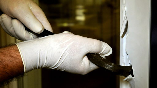 Eine Person mit Latex-Handschuhen versucht mit einem Brecheisen ein Fenster aufzubrechen.