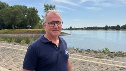 Ein Mann mit kurzen, grauem haar und einer dünnen Pioptenbrillen schaut in die Kamera, hinter ihm ist ein Fluss mit niedrigen Pegelstand zu sehen. 