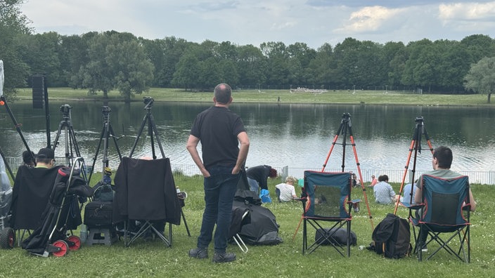 Am Rheinauensee stehen mehrere Menschen mit Kamerastativen bereit. Einige sitzen in Campinhgstühlen. 