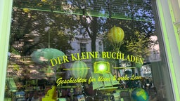 Man sieht ein Schaufenstler, darin hängen Lampenschire. In grünen buchstaben steht auf dem Glas "Der kleine Buchladen".