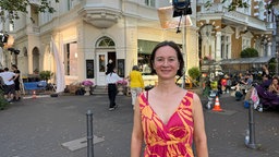 Eine circa 40-Jährige Frau steht vor einem Eckhaus., Sie hat nach hinten gekämmte, braune Haare und trägt ein Orange-rotes Kleid. 