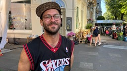 Ein Mann im Basketball-Trikot und Kappe auf dem Kopf steht vor einem Eckhaus. Er ist ca 35 Jahre alt und trägt einen Bart.